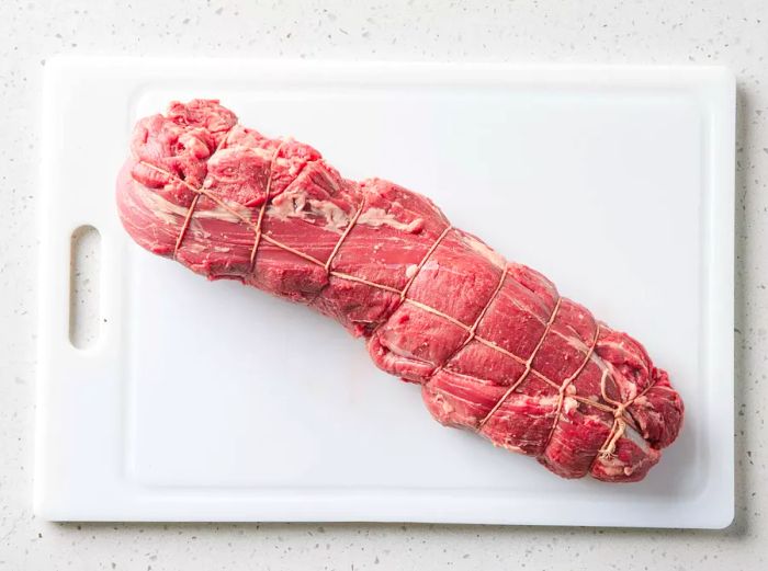 Beef tenderloin tied with string on a white cutting board.