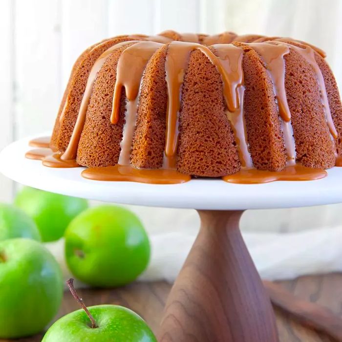 A beautiful apple pound cake displayed on a cake stand surrounded by fresh apples.