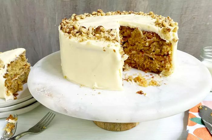 A two-tiered hummingbird cake with a slice removed, elegantly plated, sitting atop a marble and wood cake stand