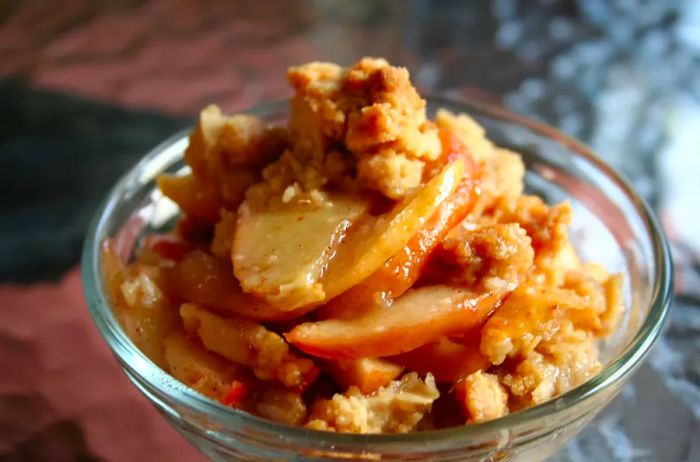 A close-up of a serving of apple crisp in a glass bowl, showcasing tender apple slices and a crunchy golden topping.