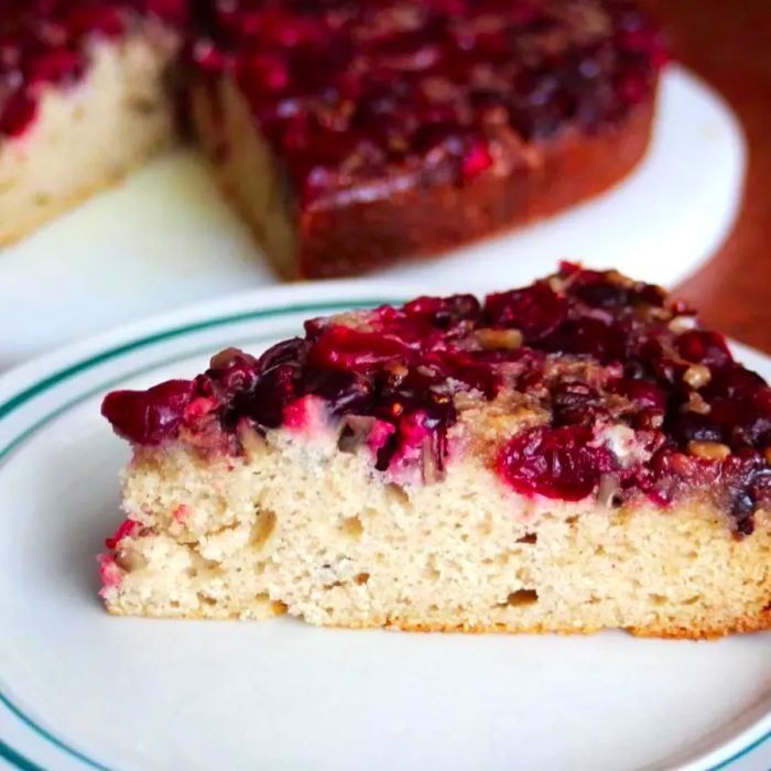 A slice of cranberry and pecan-topped coffee cake served on a white plate, with the full cake in the background.