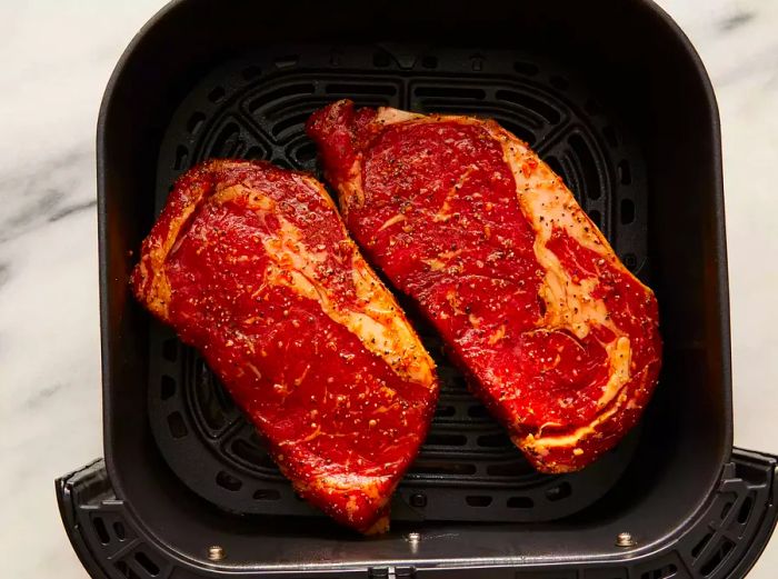 Top view of two raw steaks placed in an air fryer basket.