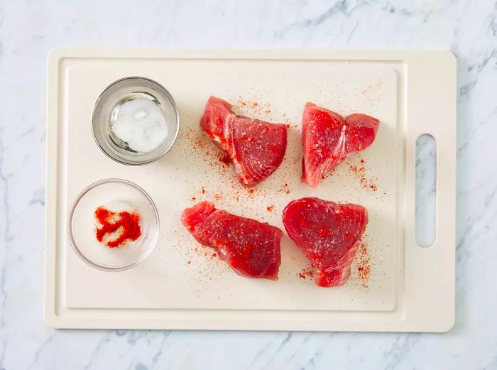 Four tuna steaks placed on a cutting board with seasonings in a bowl and sprinkled on top of the steaks