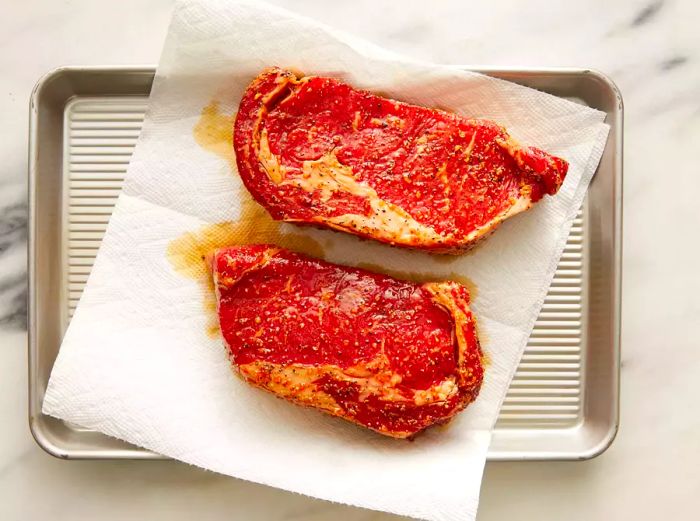 Top view of two marinated steaks resting on a paper towel-lined baking sheet.