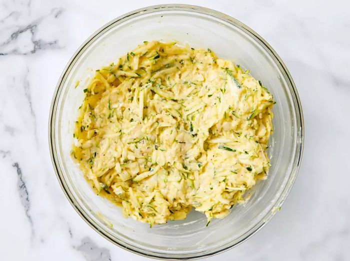 Shredded zucchini mixed with eggs, baking mix, onion, melted butter, Cheddar cheese, Parmesan cheese, rosemary, parsley, and pepper in a large bowl.