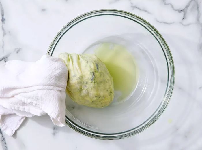 Grated zucchini being squeezed to remove excess liquid, draining into a glass bowl.