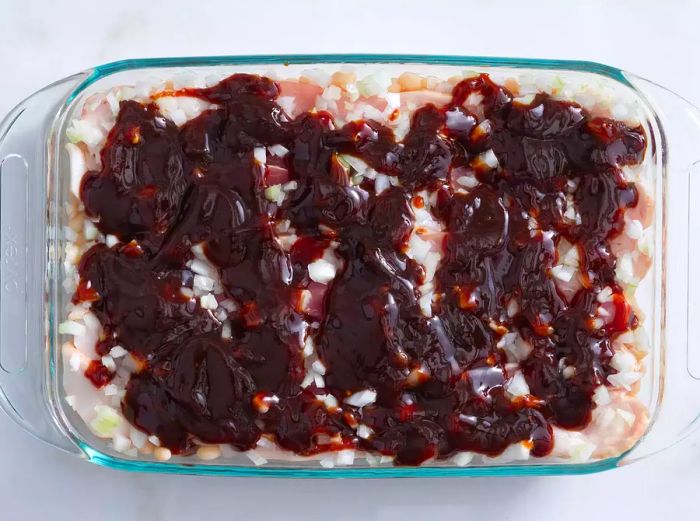 A top-down view of a baking dish layered with ingredients for Boston baked beans, topped with the sauce