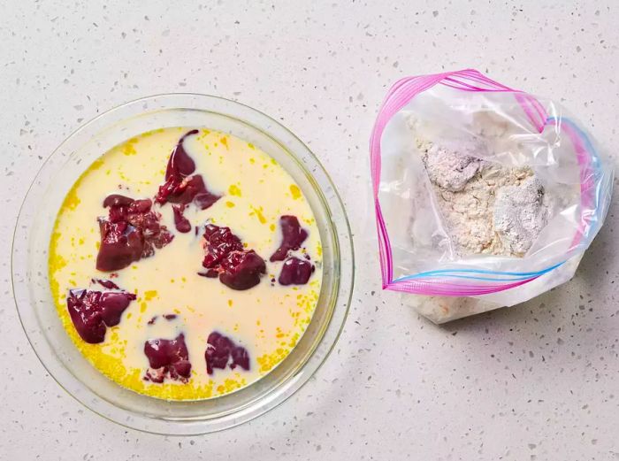 A shallow bowl of egg and milk with chicken livers, placed next to an open zip-top bag filled with seasoned flour and dredged chicken livers.