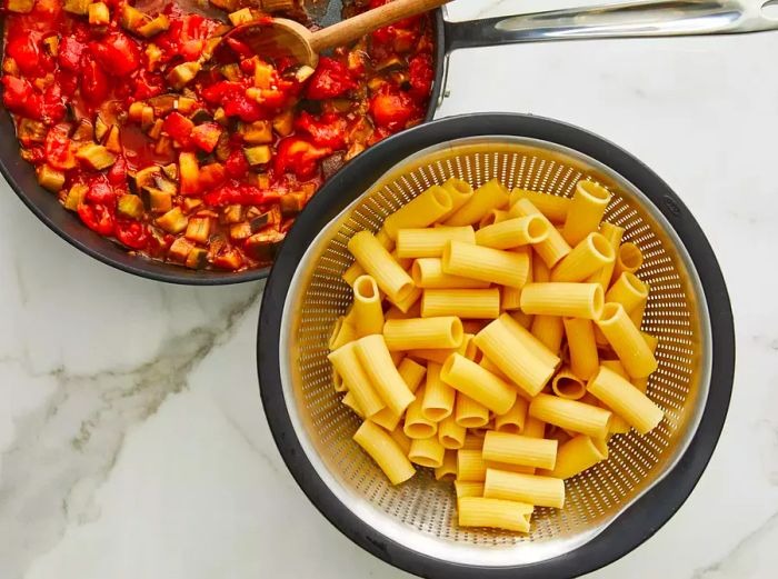 Aerial view of cooked rigatoni beside a pan of eggplant and tomato sauce