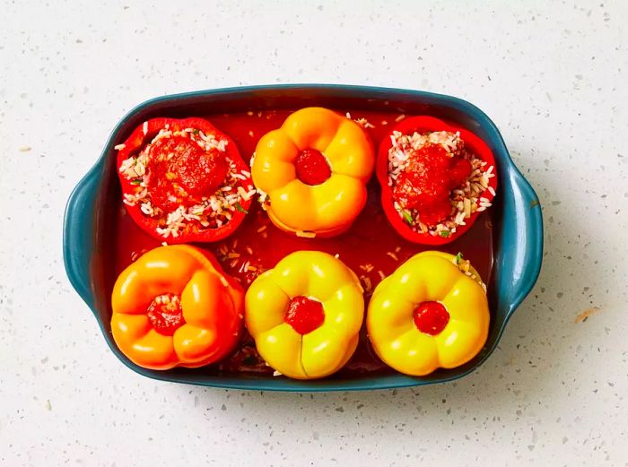 A baking dish filled with tomato sauce and sliced onions, topped with six bell peppers stuffed with a beef and rice mixture.