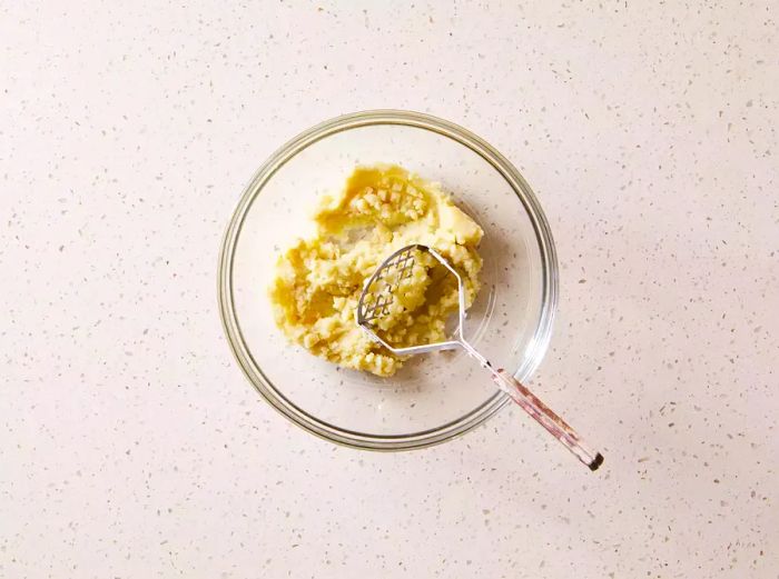 Mashed potatoes in a glass bowl with a potato masher resting beside it