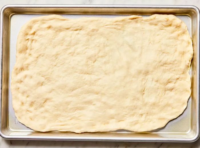 The pizza dough is stretched across a large rectangular baking sheet.