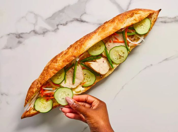 A toasted baguette, sliced in half, filled with pickled vegetables, chicken, and cucumber