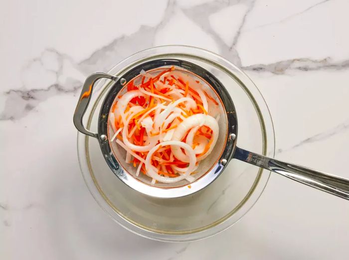 A metal mesh strainer holding pickled onions and carrots