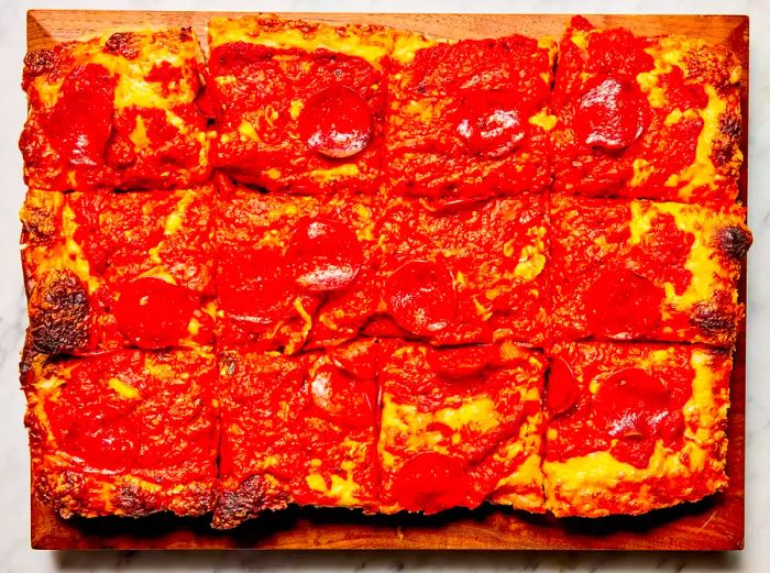 An overhead view of a slightly cooled Detroit-style pizza, cut into rectangles on a wooden cutting board.