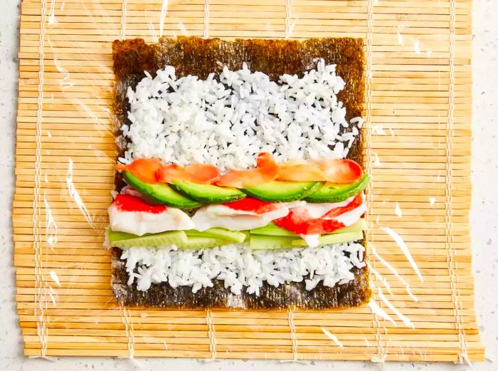 A top-down view of crabmeat, avocado, cucumber, and pickled ginger laid over rice on a nori sheet atop a bamboo sushi mat