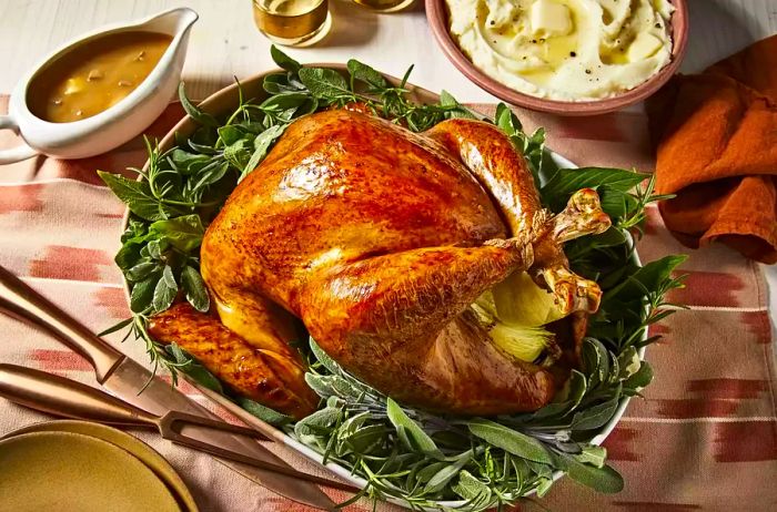 An overhead shot of a perfectly roasted whole turkey served on a platter surrounded by fresh herbs. The turkey is accompanied by mashed potatoes and gravy.