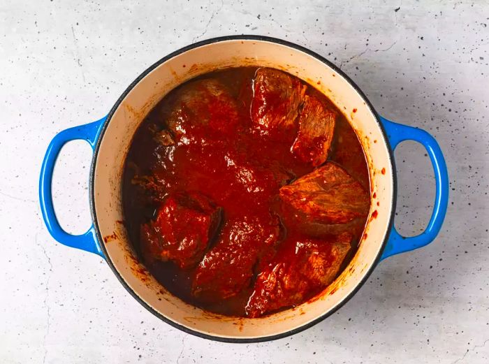 A Dutch oven with seared beef pieces covered in chili sauce