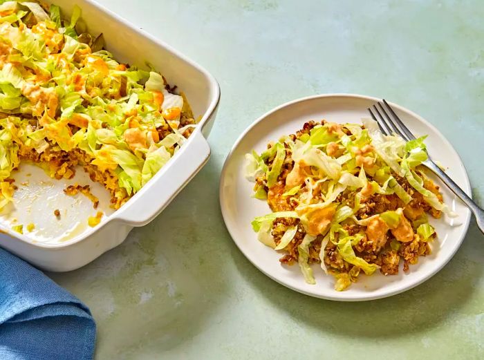 A baking dish of Big Mac casserole, with a serving plated and topped with fresh shredded lettuce and a drizzle of secret sauce