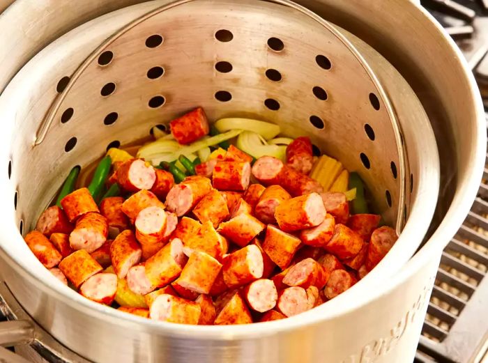 A large pot with a steamer basket, with sausage slices added on top of the vegetables.