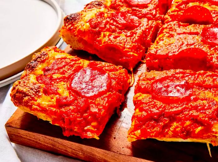 A high-angle shot of a Detroit-style pizza, sliced into squares, resting on a wooden cutting board.