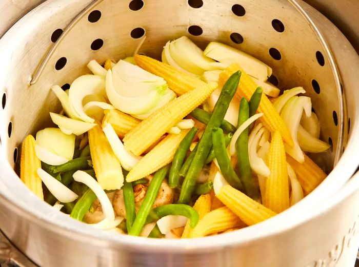 A large pot with a steamer basket containing baby corn, onions, green beans, and mushrooms added to the broth.