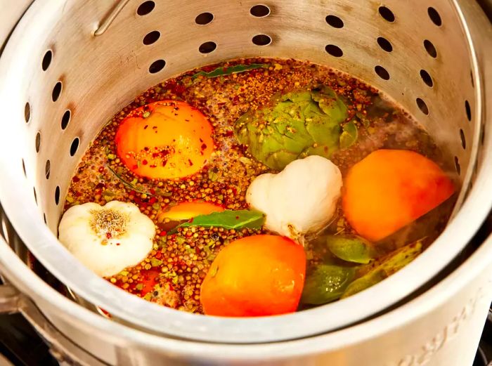 A large pot with a steamer basket filled with water, seasoning, garlic, artichokes, potatoes, lemons, and oranges