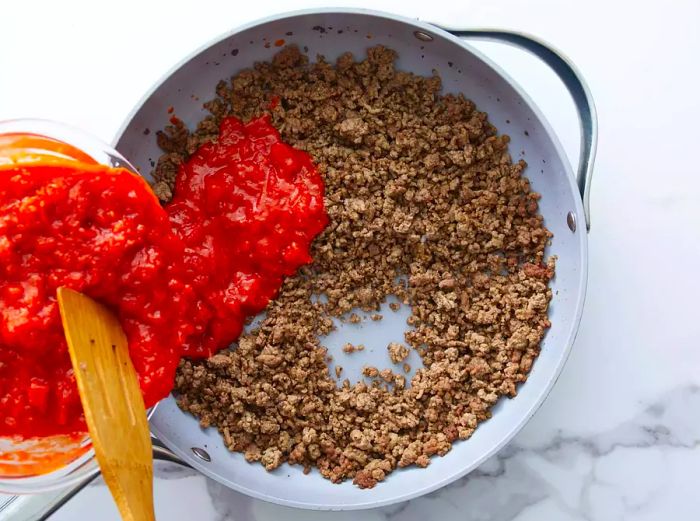 Pouring tomato sauce into the skillet with the ground beef