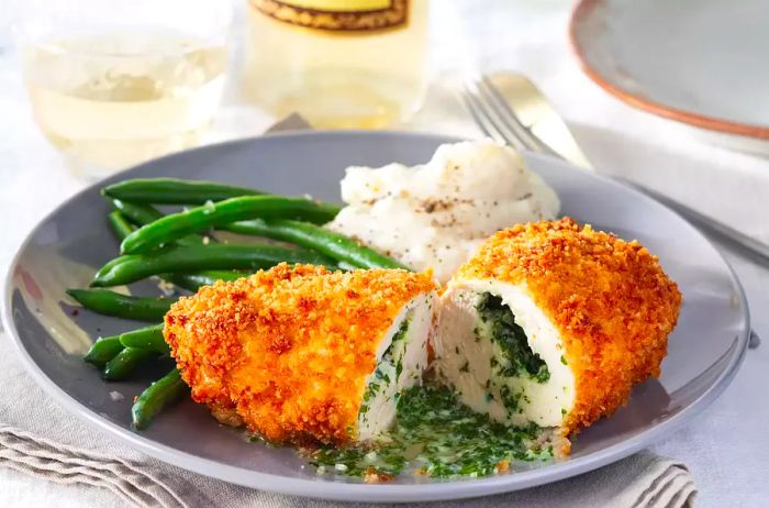 A golden-brown chicken Kiev, cut in half to reveal the herb-infused butter inside, served alongside green beans and mashed potatoes