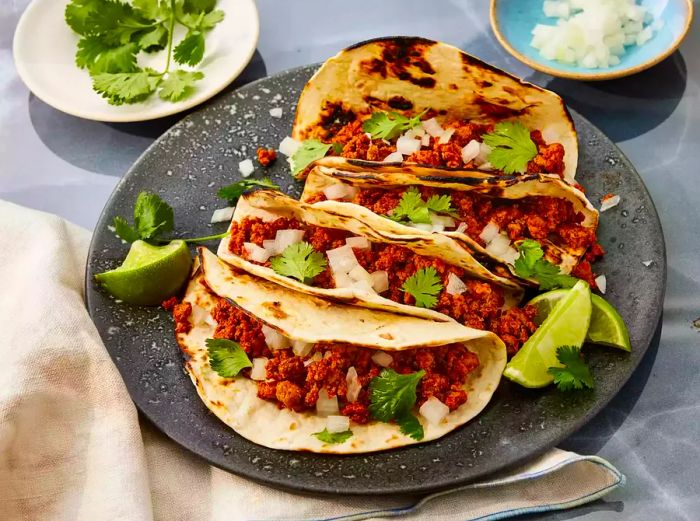 A platter with four tacos filled with ground turkey, garnished with fresh cilantro, diced onions, and lime wedges