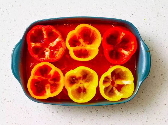 A baking dish filled with tomato sauce and sliced onions, surrounded by six hollowed bell peppers, cut side facing up.