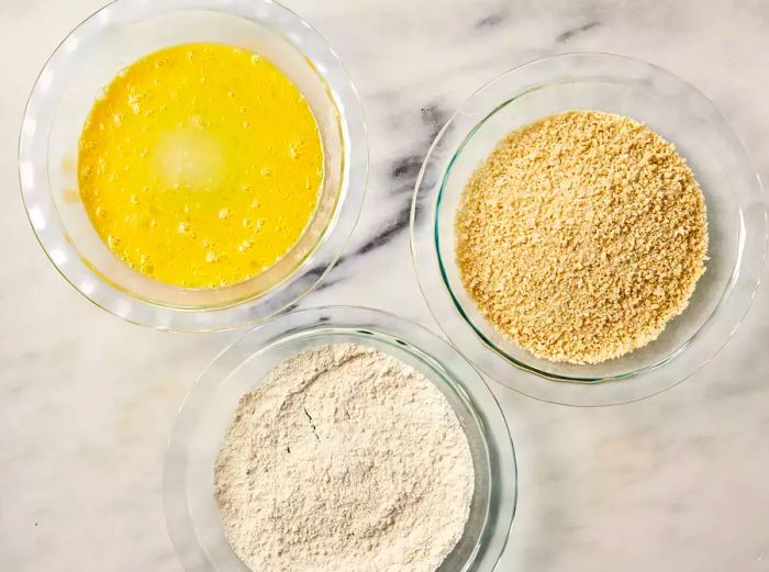 A shallow bowl with beaten eggs, a shallow bowl of seasoned flour, and a shallow bowl of breadcrumbs