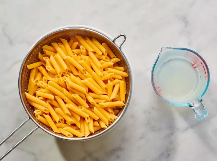 A colander filled with cooked penne pasta, accompanied by a small measuring cup of pasta water.