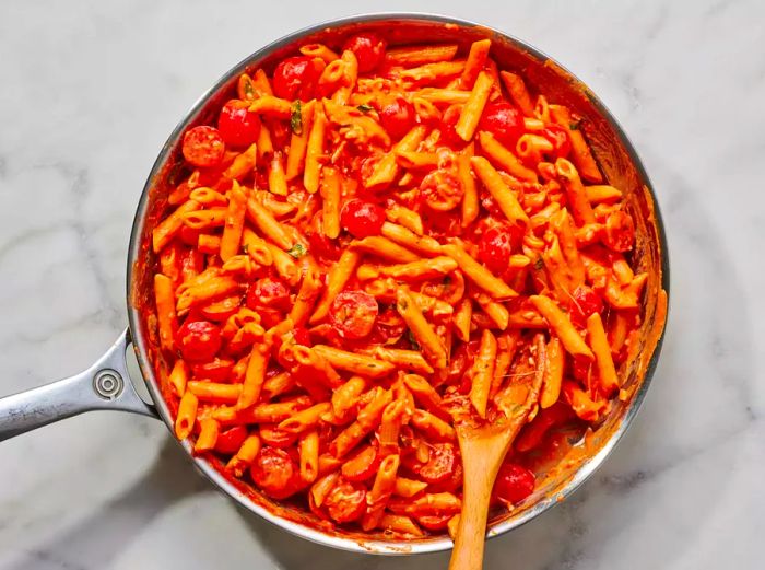 A large pan of penne pasta in creamy tomato sauce, topped with cherry tomatoes, mozzarella, and basil.