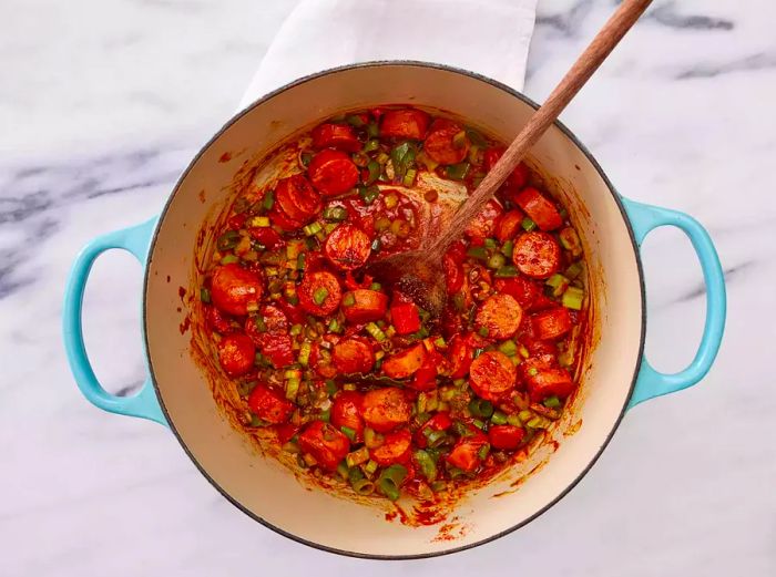 A pot filled with chopped onions, tomatoes, celery, peppers, sliced sausage, and a bay leaf.