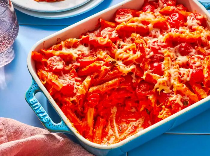 A casserole dish of creamy pasta bake with cherry tomatoes and basil, with a large portion removed for serving.