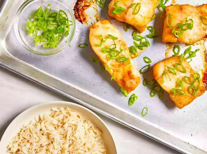 A top-down view of perfectly broiled Chilean sea bass fillets garnished with sliced green onions and served with steamed rice.