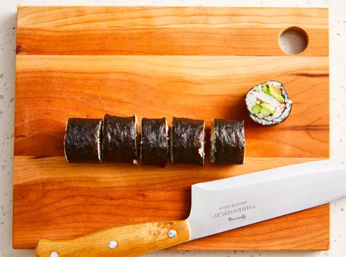 An overhead shot of sushi rolls arranged on a cutting board with a chef’s knife nearby