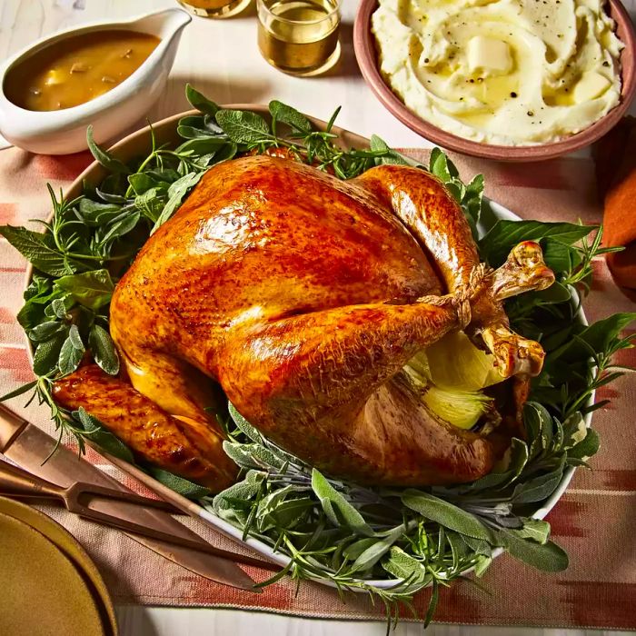 A close-up of a golden roasted turkey on a platter, garnished with fresh herbs, accompanied by gravy and mashed potatoes.