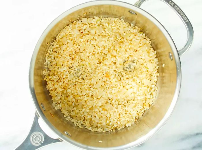 A top-down view of rice being added to a deep saucepan with sautéed onions.