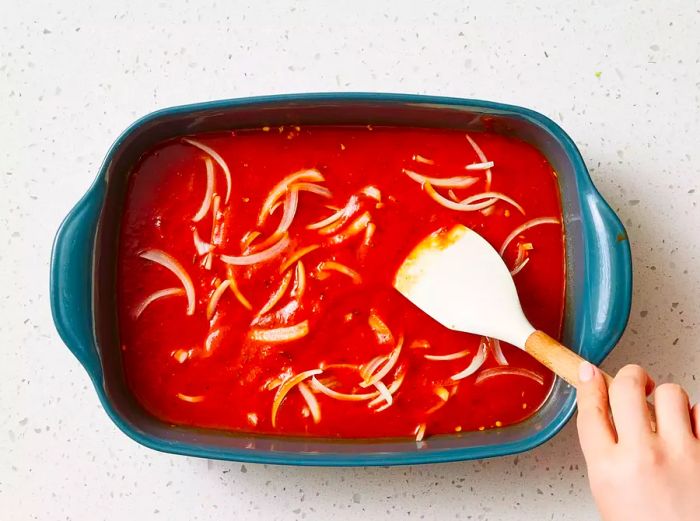 A baking dish with tomato sauce, sliced onions, and beef broth being mixed together with a rubber spatula.