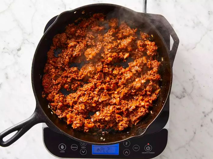 Top view of ground beef cooking in a skillet, with tomato sauce, wine, and herbs stirred in.