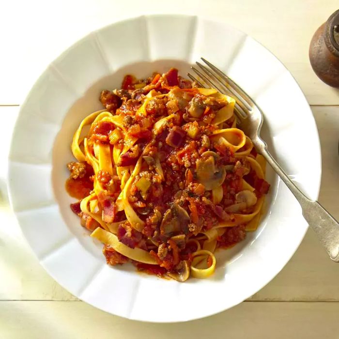 Close-up of Bolognese Sauce served with pasta in a white bowl, garnished with a fork beside it.