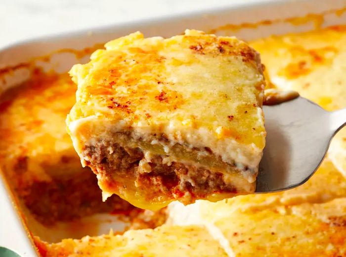 Mid-angle view of a casserole dish filled with moussaka, with a serving being lifted out.