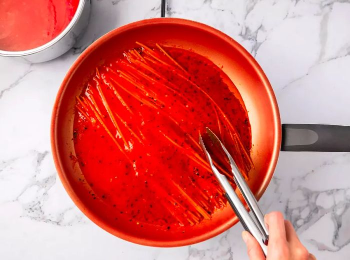 A person tossing spaghetti noodles in a hot skillet.