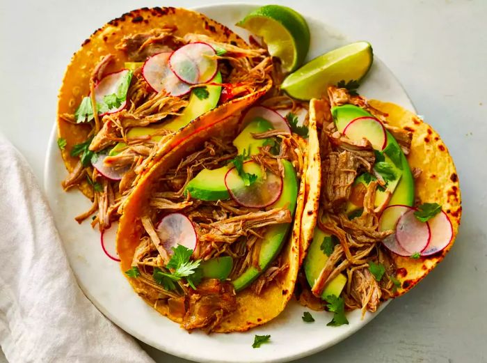 A close-up view of three tortillas filled with tender pork carnitas, topped with avocado, radishes, and cilantro, served with wedges of lime.