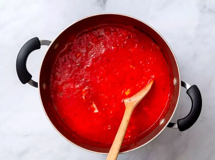 A large pot filled with crushed tomatoes, water, chopped clams, olive oil, onion, red pepper flakes, garlic, oregano, parsley, and salt, being stirred with a wooden spoon