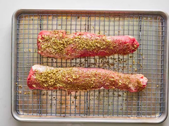 Two seasoned pork tenderloins resting on a wire rack over a baking sheet