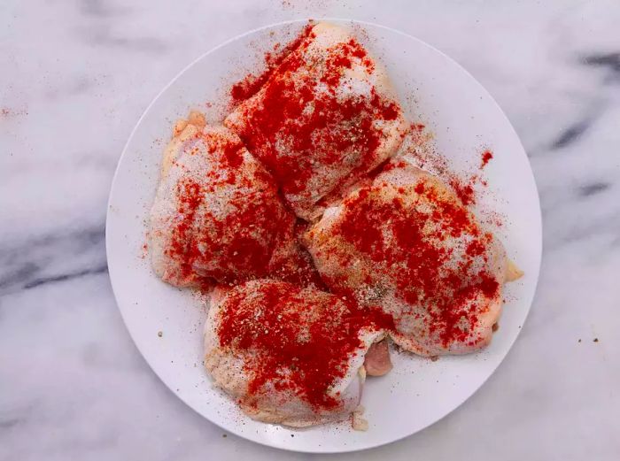 A plate holding four chicken thighs, evenly coated with paprika, salt, and pepper.