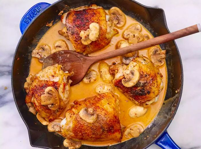 A skillet with chicken thighs and sliced mushrooms in a rich cream sauce.